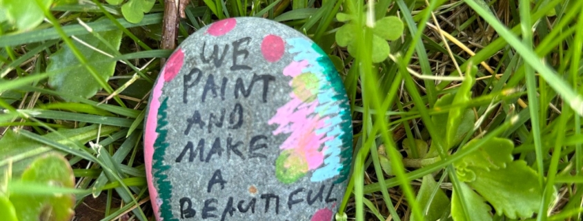 Painted rock resting in the grass, which reads "We paint and make a beautiful world - Gabe, 16"