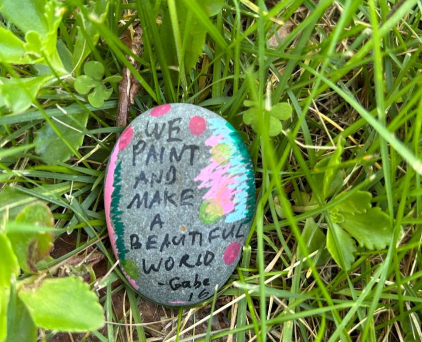 Painted rock resting in the grass, which reads "We paint and make a beautiful world - Gabe, 16"